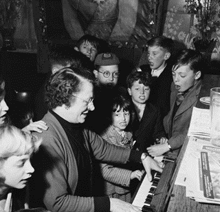 Jamaica Road, pub landlady 'Auntie Doll' (Mrs Bill Barritt) accompanies a group of singing children on the piano at the Royal George pub in Rotherhithe, 1954.  X..png
